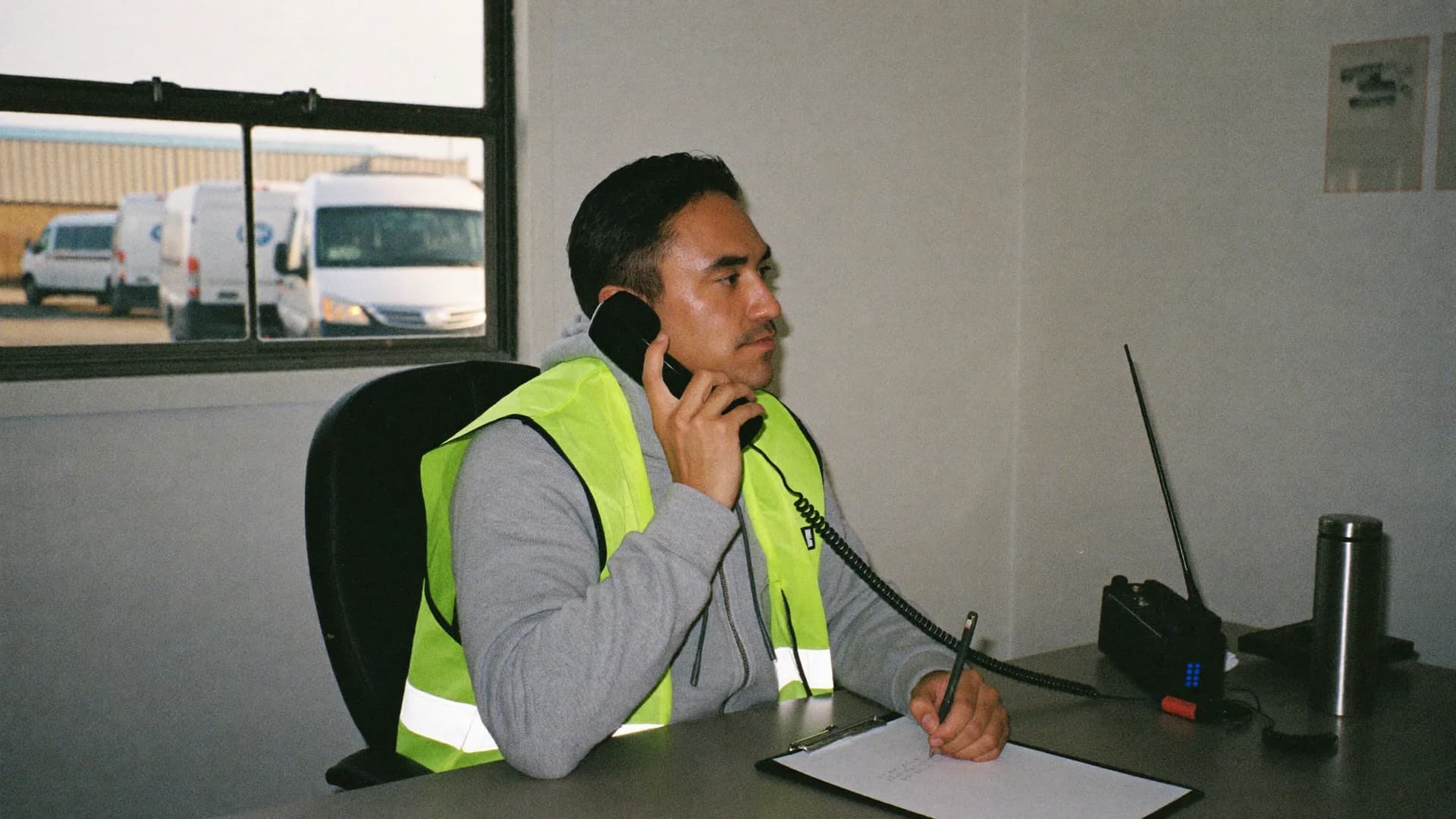 A dispatcher at his desk with the phone to his ear, the transport vans parked outside the window